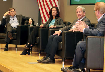 Four people sit in chairs on a stage in front of an American flag.